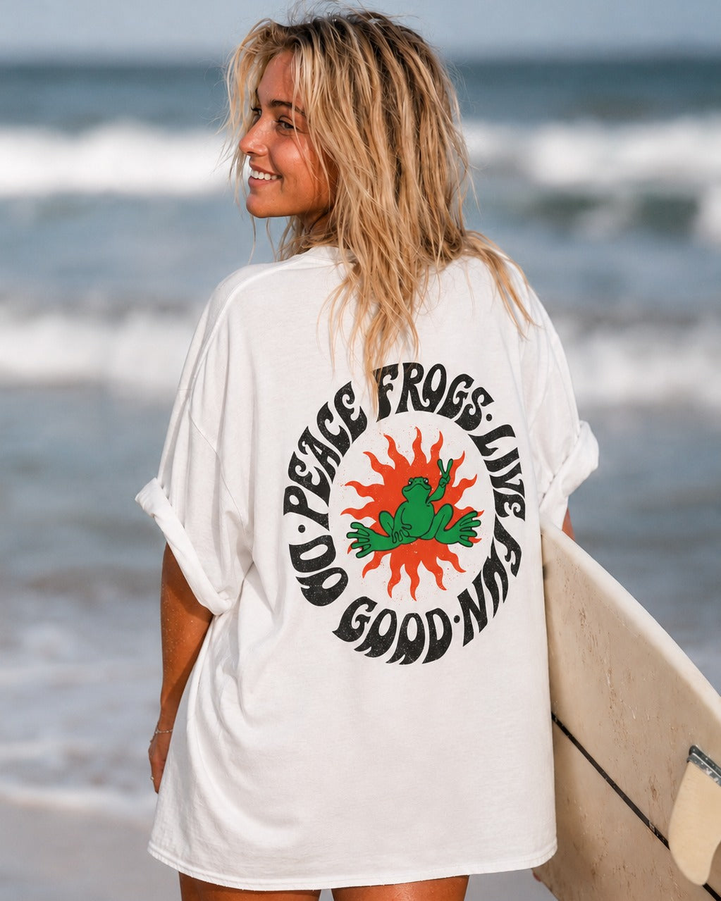 Woman on the beach holding a surfboard, wearing the white Peace Frog Sun tee with its round frog-and-sun back graphic